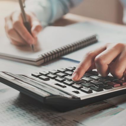 Close-up of a person using a calculator with one hand while writing in a spiral notebook with the other hand.