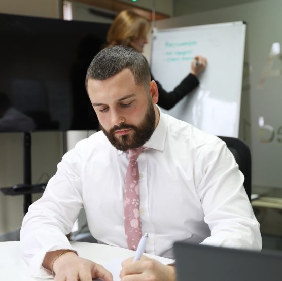 Male recruitment consultant writing at a desk with female recruiter behind him writing on a white board